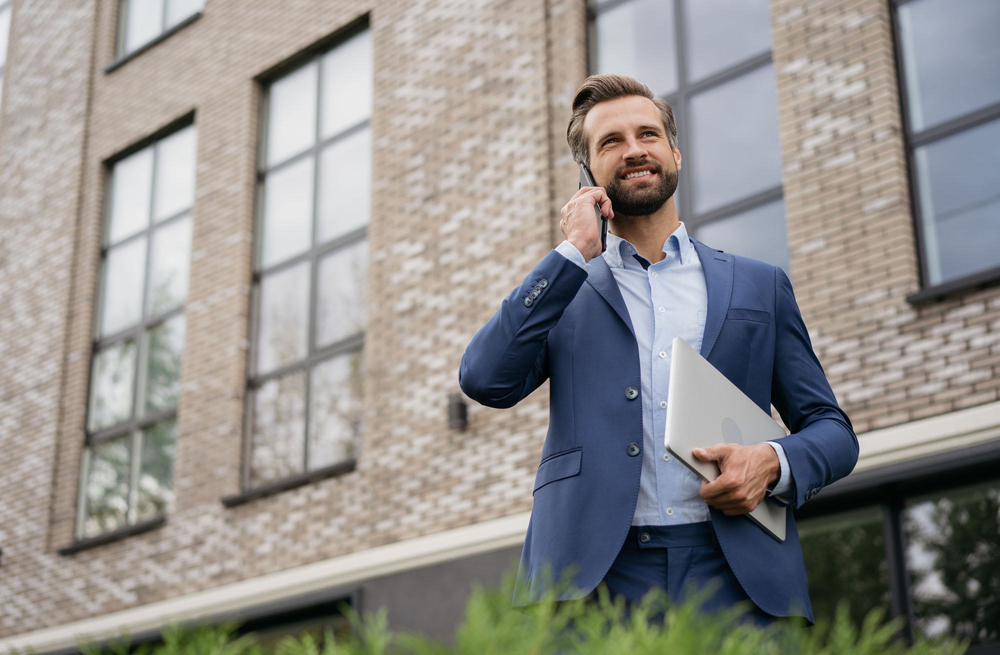 Photo of a white man wearing a blue business suit smiling as he talks on a cellphone and holds his closed laptop in his other hand. He stands outside in front of a nondescript brick building.