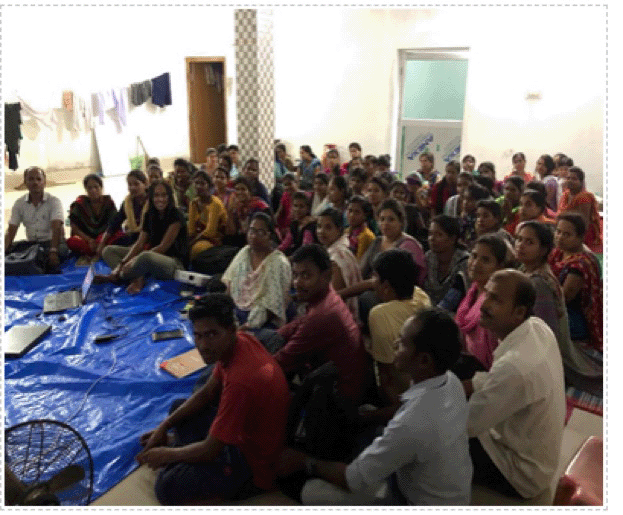 Photo of a large group of dark-skinned Indian people gathered and seated in a large, organized cluster around a blue tarp. Yilma, a dark skinned woman, is seated at the front on the blue tarp.