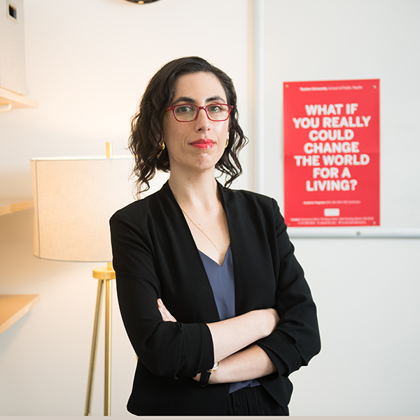 Photo of Dr. Sarah Lipson, a BU assistant professor of Health Law, with a red sign behind her that says “What if you really could change the world for a living?”