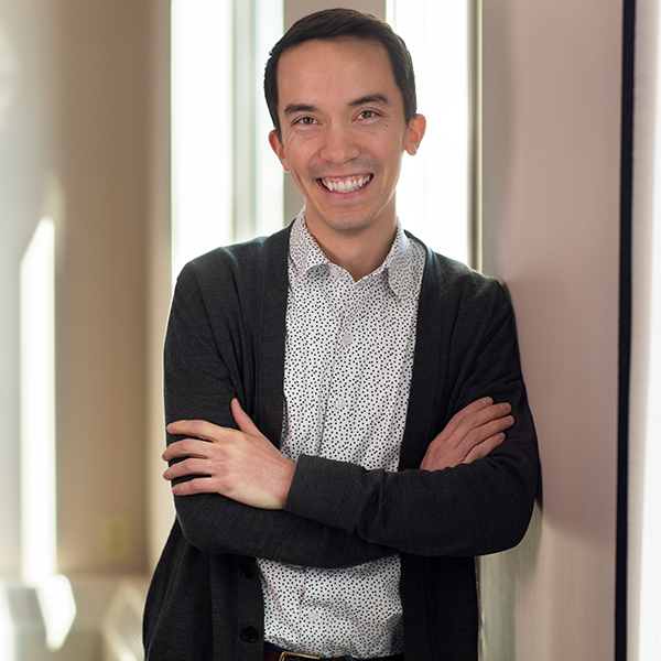 A portrait photo of Tyler Sit, a young asian american man standing wearing a light blue collared shirt and black cardigan stands with arms crossed as he smiles at the camera
