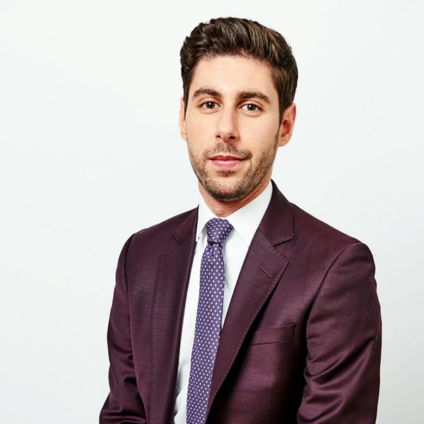 Photo of Ben Levine, a young, caucasian appearing man wearing a dark burgundy suit, white collared shirt, and lavender tie. He smiles lightly as he looks to the camera and poses.