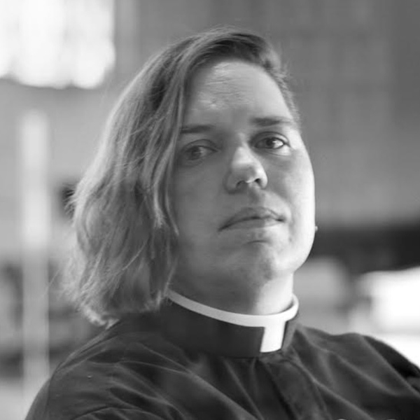 photo of Kori Pacyniak looking sternly at the camera as they wear the traditional black Catholic priest garbs.
