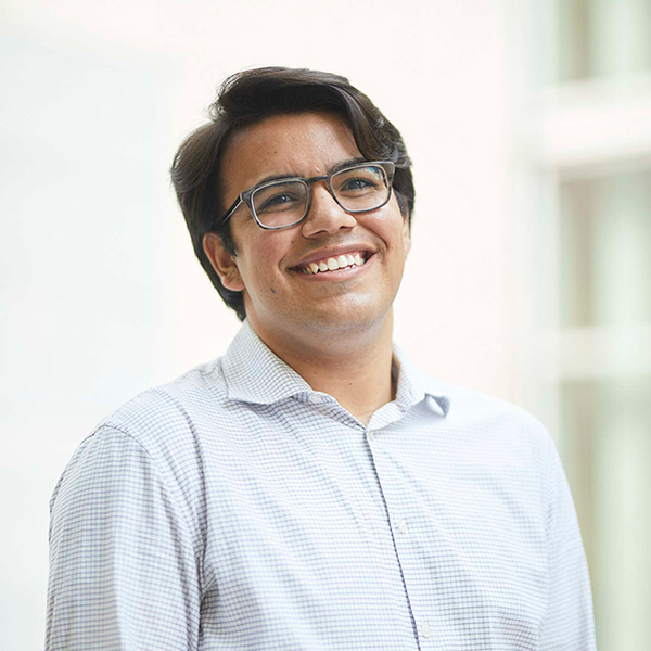 Photo of Julian Burlando-Salazar, a young Latino person wearing glasses and light blue collared shirt. He poses and smiles for the camera