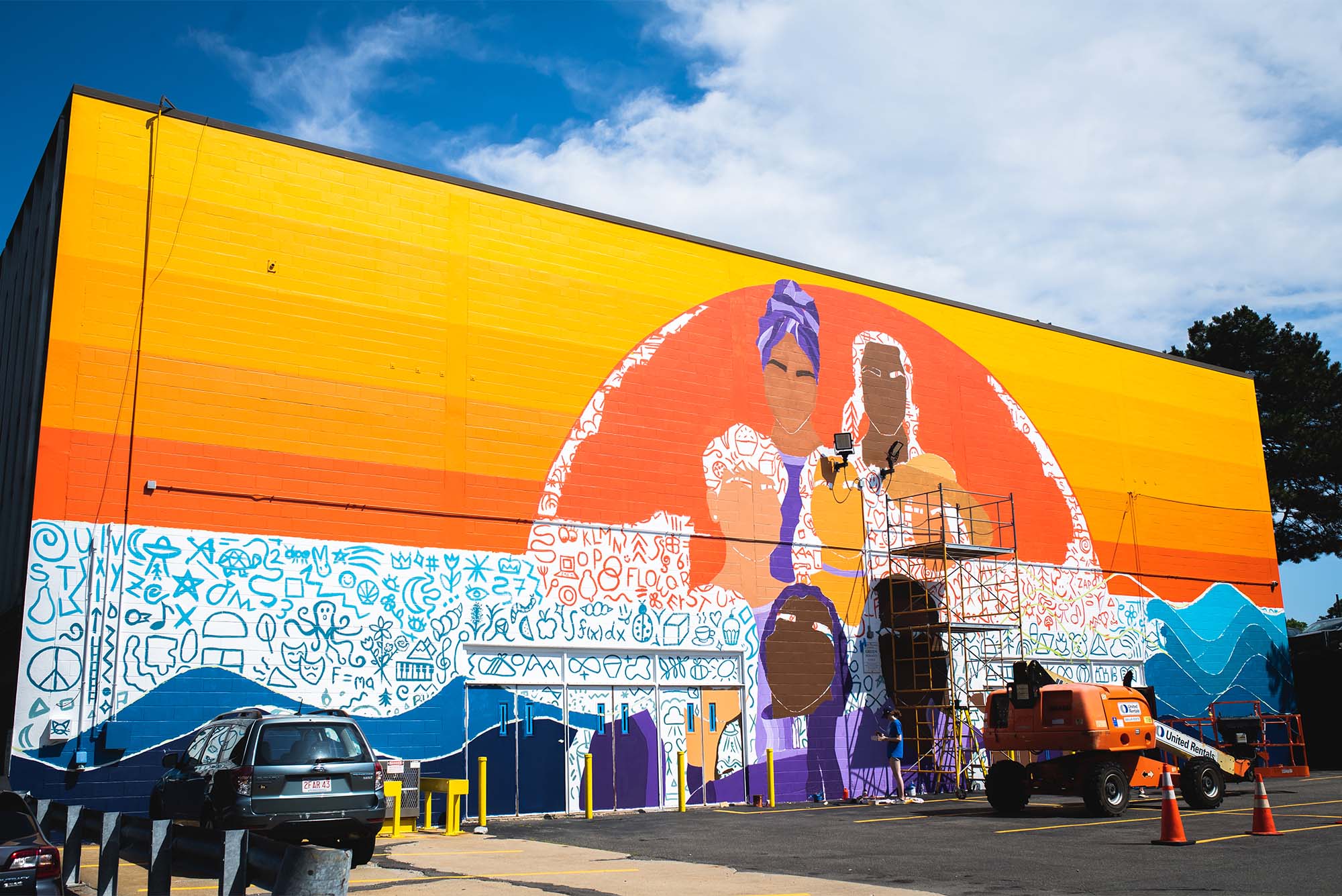 A mural being painted outside of the BUA gym in Boston, MA on July 19, 2022. The mural is orange with the beginnings of faces being painted in the center of the wall. There are smaller characters and symbols in blue on a white background below the orange expanse. Recent BUA graduate Sitarah Lakhani conceived of this project in her sophomore year at BUA. The mural is a collaboration between Sitarah, BUA, the BU Arts Initiative, and artist Amanda Hill. Recent graduate Sitarah Lakhani ’22 conceived of this project in her sophomore year at BUA, and her vision is finally coming to fruition this summer. The mural is a collaboration between Sitarah, BUA, the BU Arts Initiative, and artist Amanda Hill. The wall outside the BUA gym has been primed, and painting starts today! We’ve invited members of the BUA and BU community to participate by signing up for slots to help paint throughout the summer. The mural project is being funded privately through grants secured by Sitarah. Photo by Jackie Ricciardi for Boston University