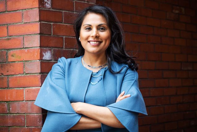 Portrait of Dr. Nahid Bhadelia leaning against a brick building. A tan woman with long black hair wearing a light blu pantsuit ensemble leans against a brick wall