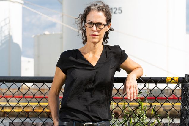 Photo of School of Public Health professor Madeleine Scammell leaning back against a black wired fence. A woman with curly brown hair, glasses, and a black button-down blouse poses for the camera.