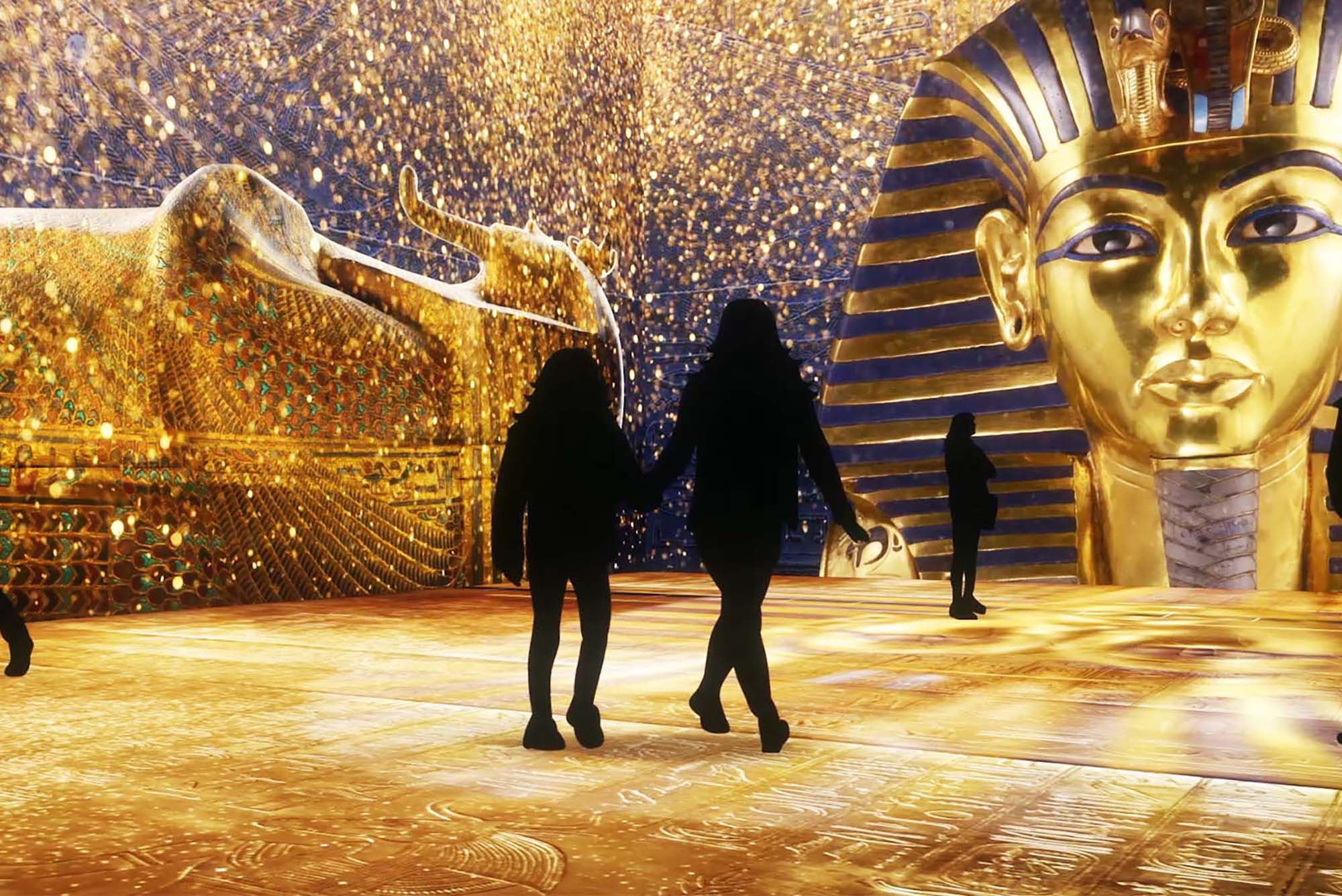 Photo of visitors stepping into an immersive gallery of golden treasure at the Beyond King Tut exhibit. Two people shown as shadows walk through a large golden exhibit. Large, golden King Tut head is seen at the entrance.