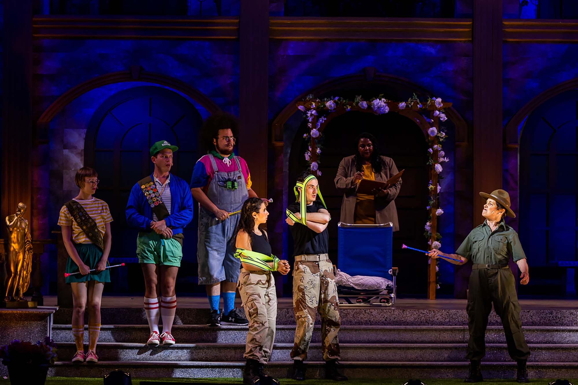 Photo of the company of Much Ado About Nothing during a dress rehersal. On stage, seven people people are seven. At right, a person dressed as a park ranger looks upset and shakes a baton at young people dresses in camoflauge pants and green vests at right. Behind those folks, what appear to be cub scouts are seen. At the back, underneath a trellis, a Black woman reads from a binder.