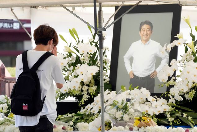 photo of a young Japanese person offering flowers and bowing their head in prayer in front of a black-framed photo of former Prime Minister Shinzo Abe, at Zojoji temple prior to his funeral wake Monday, July 11, 2022, in Tokyo. In the photo, Abe wears a white button down shirt, black pants, and smiles with his hands on his hips. Large, white flower displays are seen around the photo and on the table in front. The saddened young person wears a white shirt and a black backpack and has short light brown hair.