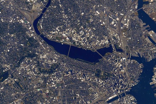 Photo of BU, and the surrounding Boston area, taken from the International Space Station. The Charles River is deep dark blue, and the streets and surrounding houses are mostly gray in color. You can see Nickerson Field and the BU bridge in the image.