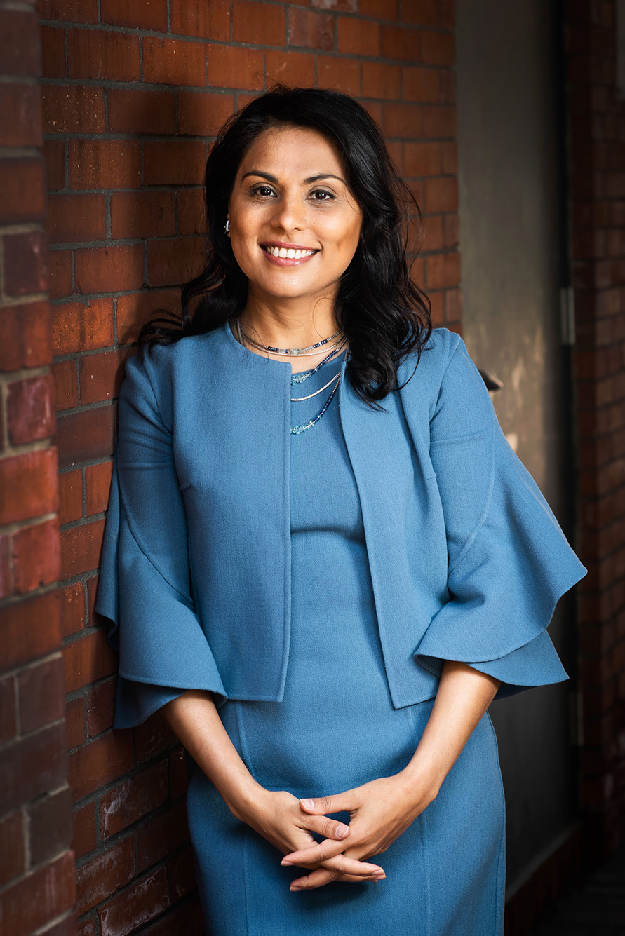 Portrait of Dr. Nahid Bhadelia leaning against a brick building. A tan woman with long black hair wearing a light blu pantsuit ensemble leans against a brick wall