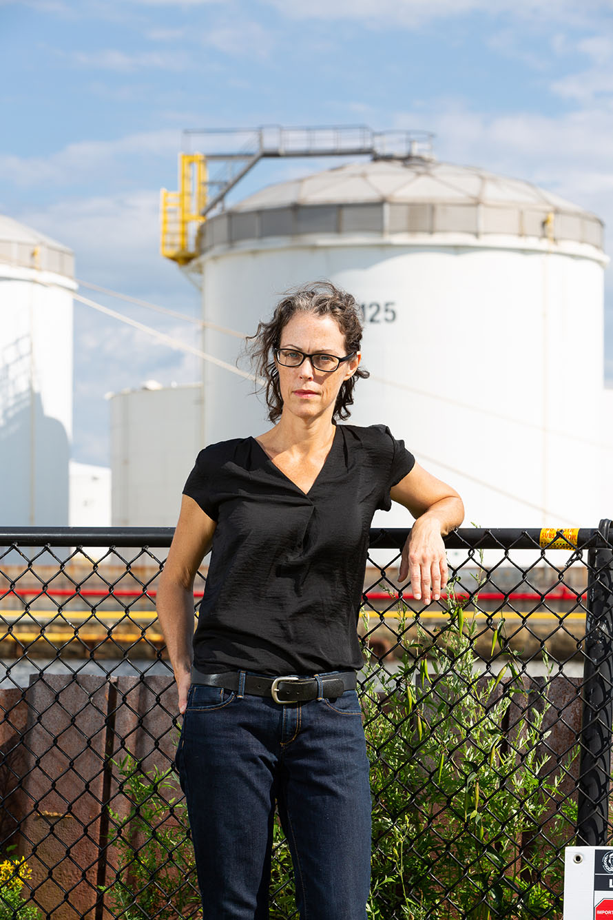 Photo of School of Public Health professor Madeleine Scammell leaning back against a black wired fence. A woman with curly brown hair, glasses, and a black button-down blouse poses for the camera.