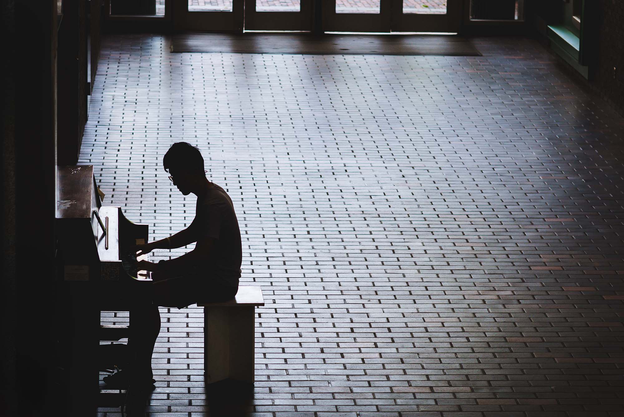 Photo, taken slightly from above, of Kangbo Li (ENG 22) practicing the piano in the GSU on June 29, 2022. Li is seen in silhouette, the piano is almost completely blacked out in shadow. The photo is essentially black and white. The bricks of the GSU reflect a silvery color.
