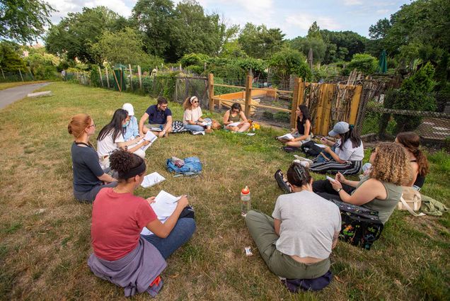 photo of a group of students in MET’s Urban Agricultural class sitting in a circle for a lecture before taking a tour through the Fenway Victory Gardens July 7, 2022. The students wear summer-y clothing and sit on a patch of grass just outside of a garden with a wooden and wire fence.