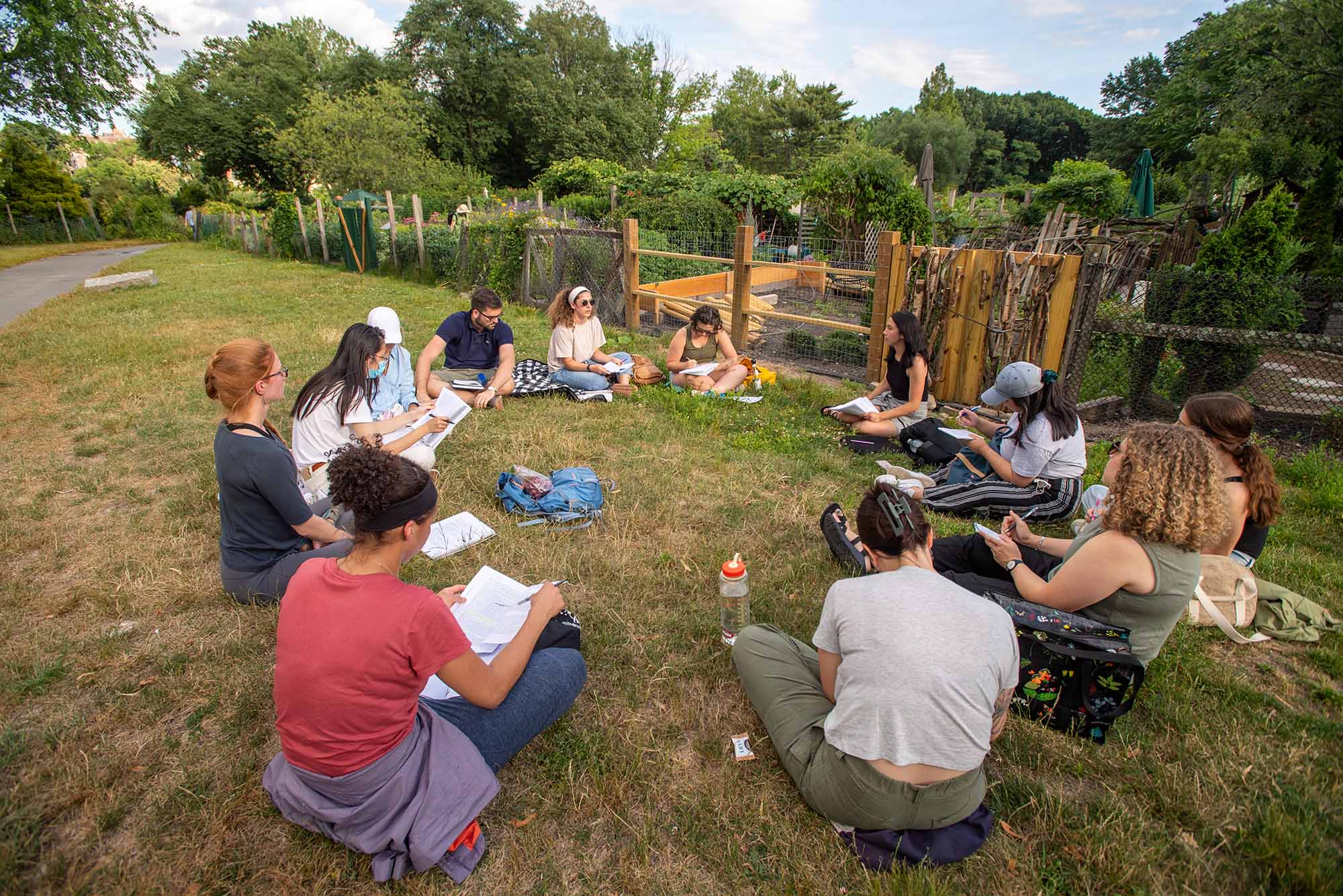 photo of a group of students in MET’s Urban Agricultural class sitting in a circle for a lecture before taking a tour through the Fenway Victory Gardens July 7, 2022. The students wear summer-y clothing and sit on a patch of grass just outside of a garden with a wooden and wire fence.