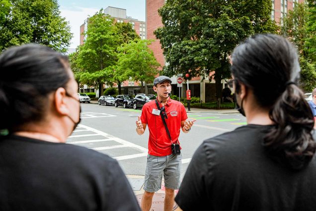 Photo of student tour guide Dante Pettinato (CAS'25) talking about on-campus residence options while walking on Bay State Road on July 8th. He wears a gray scallycap, a red Boston University polo shirt, and gray shorts. He speaks into a microphone, and the speaker hangs at his side. The shoulders of masked tour attendees are seen in the foreground. The trees and cars parked on Bay State Road are seen behind him.