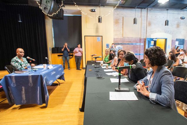 Photo of Micaelah Morrill of innovate@BU, far right, discusses food insecurity on college campuses and solutions to the problem while Congresswoman Ayanna Pressley, left, listens during a WH Hunger Summit District Convening in East Boston July 11. Morrill is a white woman with ear-length curly hair and wears a light blue blazer and clasps her hands as she speaks at the microphone. She and six other people sit at a table with a black tablecloth and mics in front of them. Pressley, who is seen slightly blurred, wears a blue, green and white blouse and sits by herself at a table with a blue tablecloth. Photographers can be seen at the other end of the room.