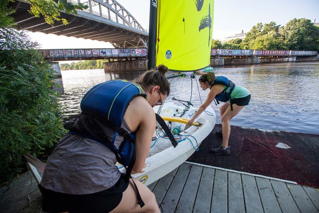 Photo of students in BU’s Learn to Sail class, including Chelsea Mattioda (LAW’22), left, and Anna Carr, music accompanist at CFA, launching from the boating pavilion for lessons not he Charles River July 12. Mattioda and Carr both wear blue and green life jackets. Carr takes the front of the small, white boat (a Quest) with a bright green-yellow sail, while Mattioda, her hair in a bun, pushes from the back. The graffiti on the old rail track underneath the BU bridge is seen at left; the Charles at right.