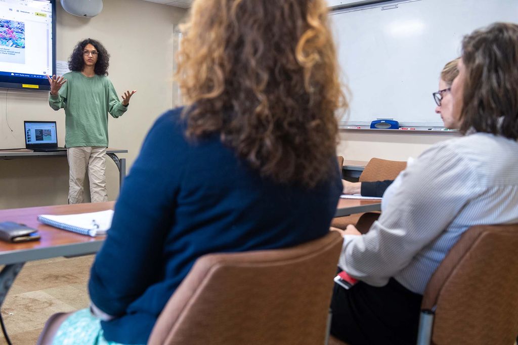Photo of high schooler Mihailo Stenovic, in the background, at left, presenting at a Shark Tank style pitch competition as part of the PopHealth 2 week summer program July 15 at MED. Mihailo has long, curly brown hair and glasses, light brown skin, and wears a green long sleeve shirt with khaki pants. Half of a slide on screen is seen behind him. At right, a panel of women are seen blurred from behind.