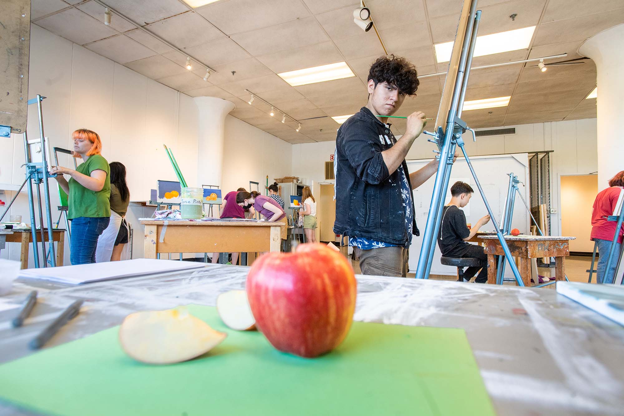 Photo of High school junior Jose Pinto of East Boston painting a still life at CFA’s Visual Arts Summer Institute (VASI) July 12. In the photo, an apple with two slices laying on the cutting board next to it, is seen blurred in the foreground. The apple is red with orange and yellow streaks. Pinto, a young person of color, has short curly hair, and looks intensely at the apple while holding a green paintbrush to the easel in front of him. Other students can be seen painting and doing other art projects in the classroom behind him.