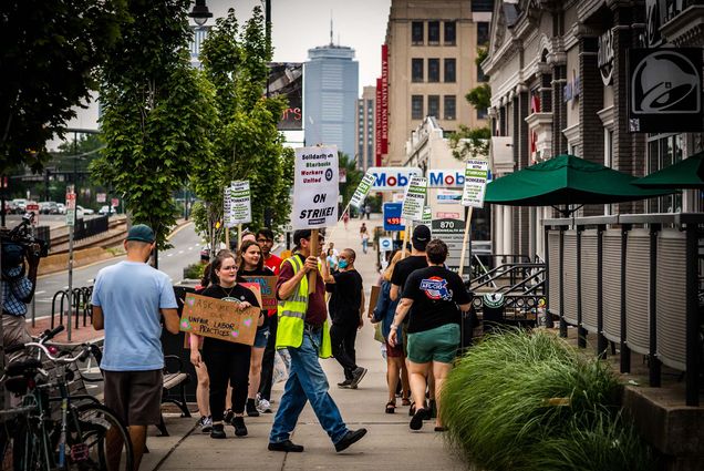 Photo of Starbucks employees picketing outside of the store location on Comm Ave. on July 18, 2022. They are a mix of genders and appear to come from diverse backgrounds; most of them are youngish people. They hold signs that say "Ask me about our unfair labor practices," "Solidarity with starbucks, workers united, on strike!," "Solidarity with Starbucks workers, fight for a living base wage of $25/hr before tips!" The prudential center is seen in the background, as well as the trees lining comm ave and the thurman center.