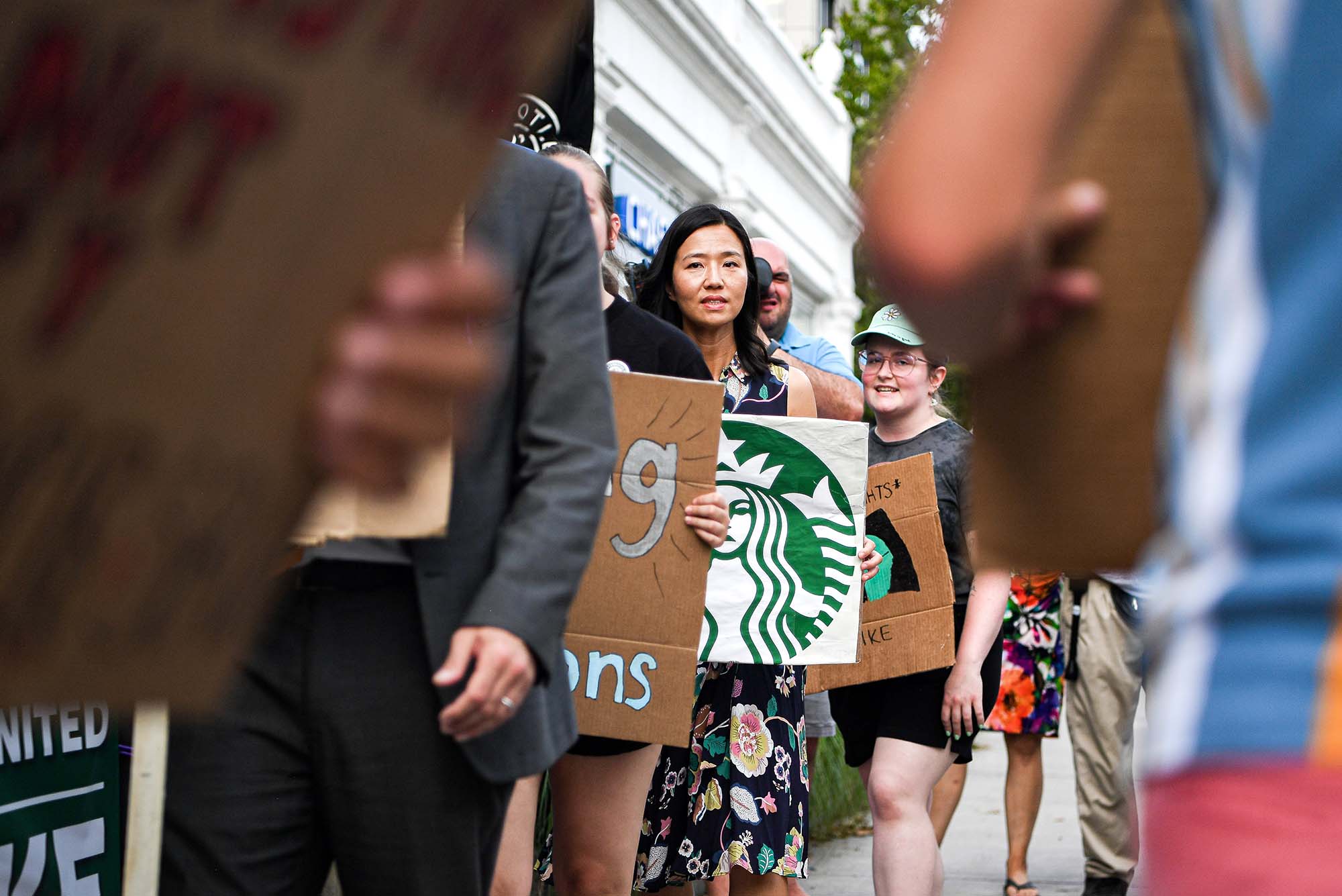 Photo of Michelle Wu, Mayor of City of Boston joining the line of protestors outside the Starbucks along Comm Ave. She wears a black dress with a floral pattern an olds a white sign that has the Starbucks logo altered so that the mermaid is frowning. Wu is a middle aged AAPI woman with shoulder-length black hair. In the foreground, and in focus, other protestors holding cardboard signs are seen. Photo taken July, 25th by Ziyu (Julian) Zhu (CGS'23)