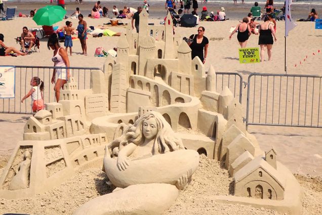 Photo of an elaborate sand castle from the 2013 festival featured a mermaid holding a shark or fish in front of a winding castle. The sculpture is fenced off and beach goers can be seen in the background.