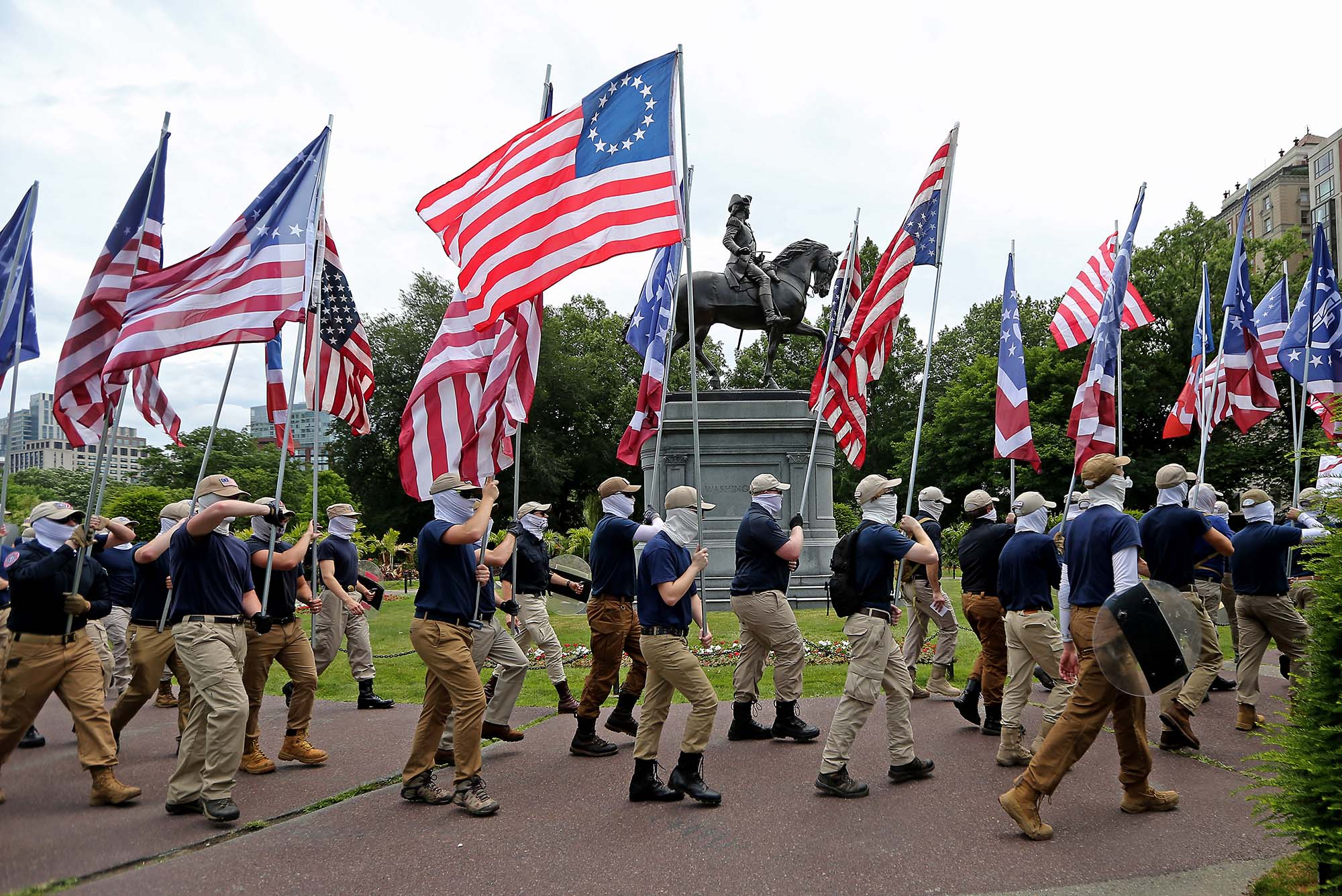 Photo of the white supremacist group, The Patriot Front marching thru the city of Boston on July 2, 2022. They walk by the statue in Boston Public Gardens holding Betsy-Ross-style flags. Their faces are covered by white-gray neck gaiters, tan baseball caps, khaki pants and blue polos. Many wear sunglasses so their face is completely hidden. There's about two dozen pictured.