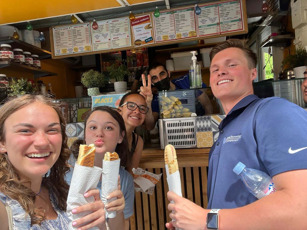 Photo of four students in the SHA Paris marketing program holding baguette sandwiches and posing with a street vendor who wears a mask and makes the peace sign with his hands. Three of the students, two women, one boy, are White, and one women of color is seen in the background. 