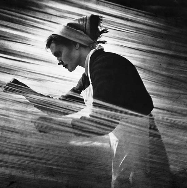 Album cover featuring a photo or painting of what looks like an early 20th century woman working or weaving. The images is in black and white, with white streaks running left to right. The woman wears her hair back in a white hanker-chief and wears a white apron.