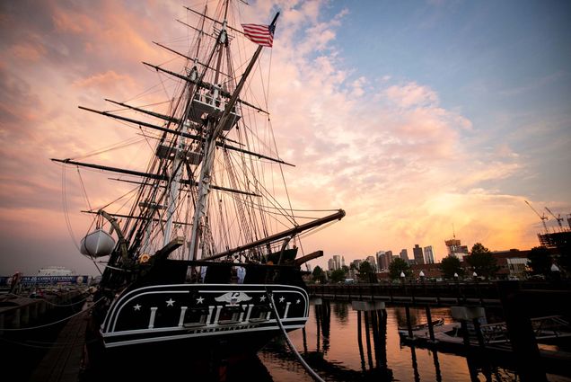 Photo of the USS Constitution, a historic warship, docked in Charlestown Navy Yard at sunset. The ship has three white masks and is painted black. The stern of the ship is decorated with six white stars and a white eagle with an American flag emblem at center. A dingy is hoisted up on its left side and an American flag flies off its right side. The sun sets behind the ship; the sky is filled with a pink fluffy cloud which is reflected in the water. Some of the Charlestown skyline is seen in the background.