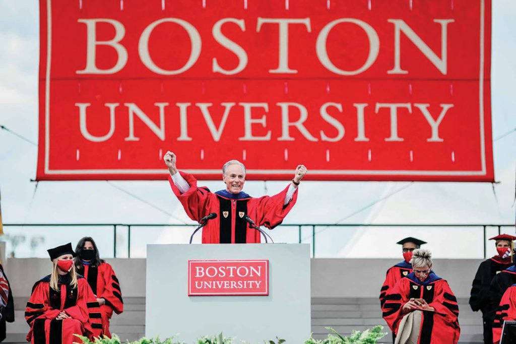 Photo of JR Hipple extending his arms outwards at the podium at graduation; he wears the ceremonial red robe and has a large Boston University sign behind him.
