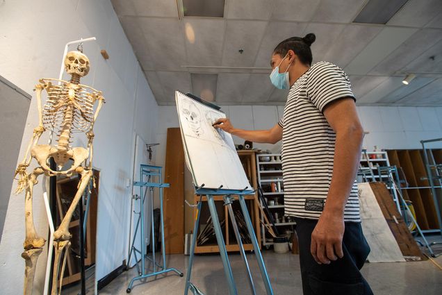 Photo of Andres Lopez, at right, wearing a white and black striped shirt and black pants, sketches a skeleton that hangs to his left, as part of Drawing 1 class held by CFA. Lopez’s sketch pad rests on a large blue easel, other easels can be seen in the classroom in the background.