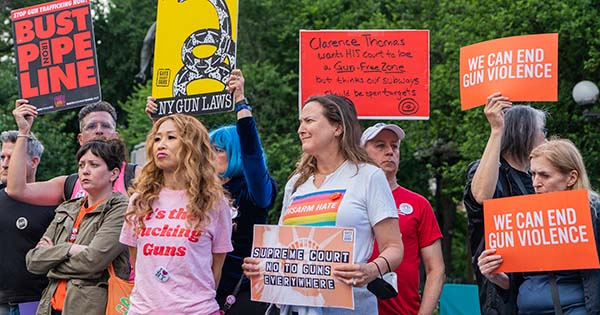 Photo of people of various ages and genders holding signs at a rally at Union Square in New York City, NY June 23, 2022 after the Supreme Court of the United States ruling on gun laws in New York. An AAPI woman with long, wavy blonde hair wears a pink shirt that says "it's the fucking guns" and a button that says "i love new york." A white woman next to her wears a shirt that says "Disarm hate" and holds a sign that reads "Supreme court no to guns everywhere." Behind her, a white man in a red t-shirt holds a sign that reads "Clarence Thomas wants HIS court to be Gun-Free-Zone but thinks our subways should be open targets." Older white women at right hold orange signs that read "we can end gun violence"