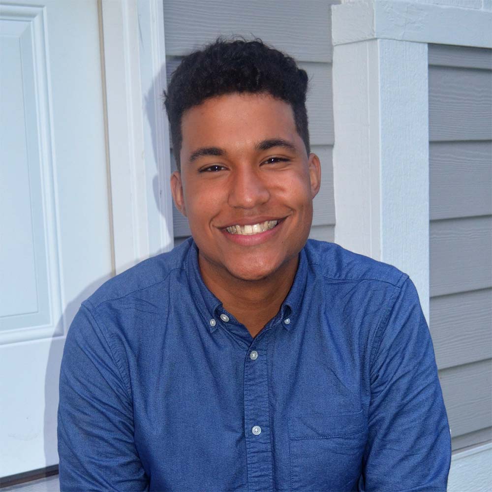 Photo of Morel, smiling, while wearing a blue button down shirt and sitting on what looks like a porch, as gray siding can be seen behind him. He is a young person of color, and has short hair, trim at the sides and curly on top.