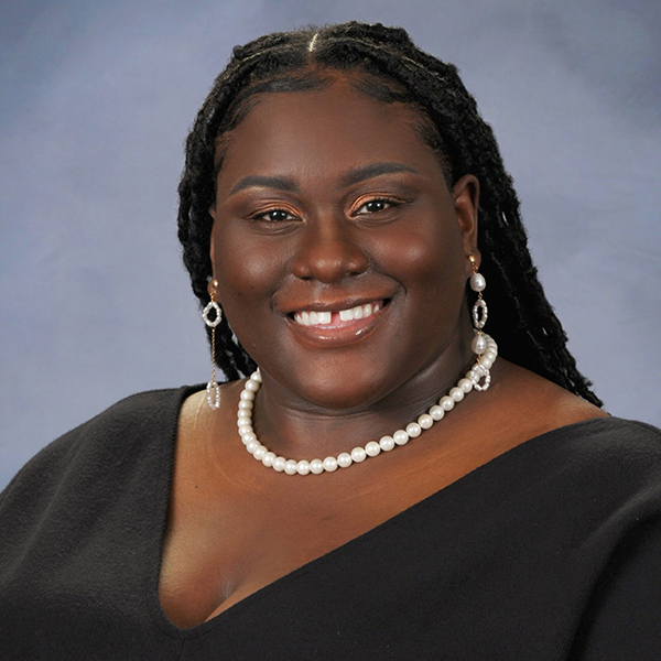 Photo of Kahjirah Taylor Harris, a young black woman with long, black Senegalese twists and wearing a black blouse and pearl necklace smiles and poses for the camera.