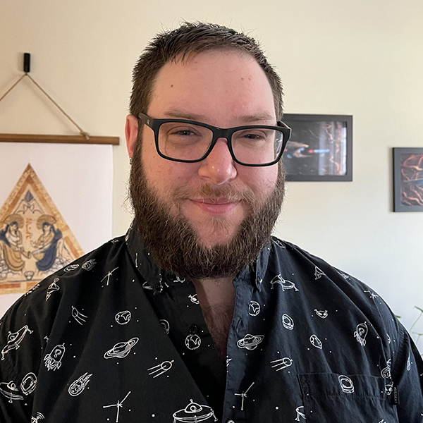 Photo of Evan McDuff, a young light skinned man with glasses and wearing a black and white patterned, collared shirt, smiling at the camera.