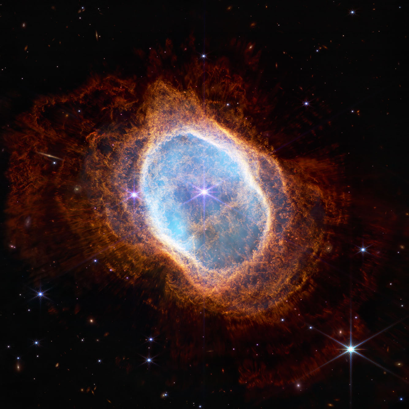 Photo of the Southern Ring Nebula. A large bright blue, transparent blob surrounded by a haze of red is shown in space.