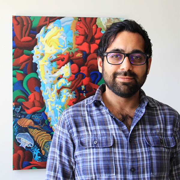 Photo of Victor Kumar. A tan man with a black beard and mustache smiles for the camera. He wears a blue plaid shirt and glasses.