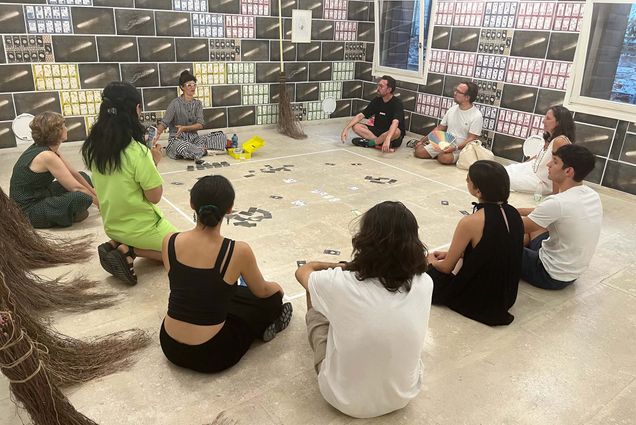 Maria Molteni (CFA’06) (center) discusses her immersive work, Counting to Infinity/Sweeping Stars (Contando All’Infinito/Stelle Scopa), at the July 30 opening for Figure, Character, Sign, an exhibition celebrating more than 20 years of BU’s Venice Studio Arts program. The wall of the room is covered with a series of printed blocks, some have 8 sets of cards on them, others have a large white streak. The group sits in a circle around Molteni, who wears a black and white striped jumpsuit and white glasses. The group is a mixture of men and women of various ages who sit on the floor. At center, cards are scattered around. At left, traditional venetian brooms, which appear to be made from hay or straw, are seen.