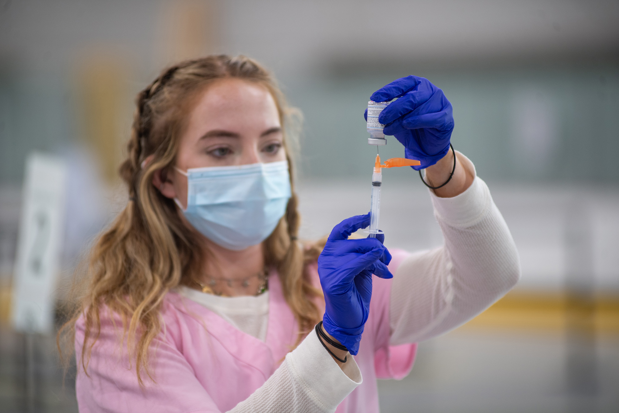 Photo of Triage nurse specialist Victoria Cunningham, in pink scrubs, blue gloves, and a blue face mask, at the Moderna vaccine clinic at FitRec May 4. She holds a vial and extracts a dose of the vaccine with a needle.