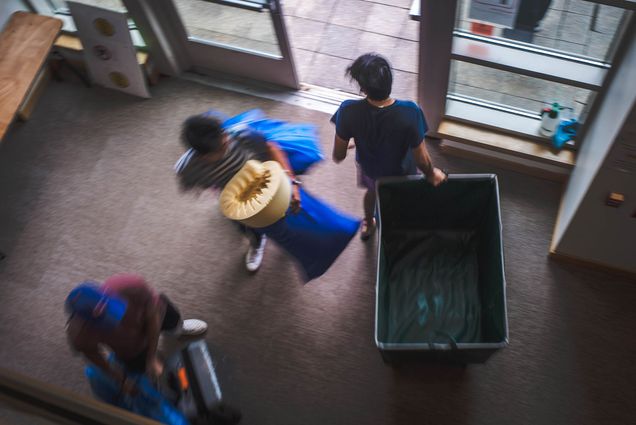 Photo of students moving back to BU's campus . A blurry overhead action shot shows students moving a large yellow cart and bed supplies as they move into a dorm building.