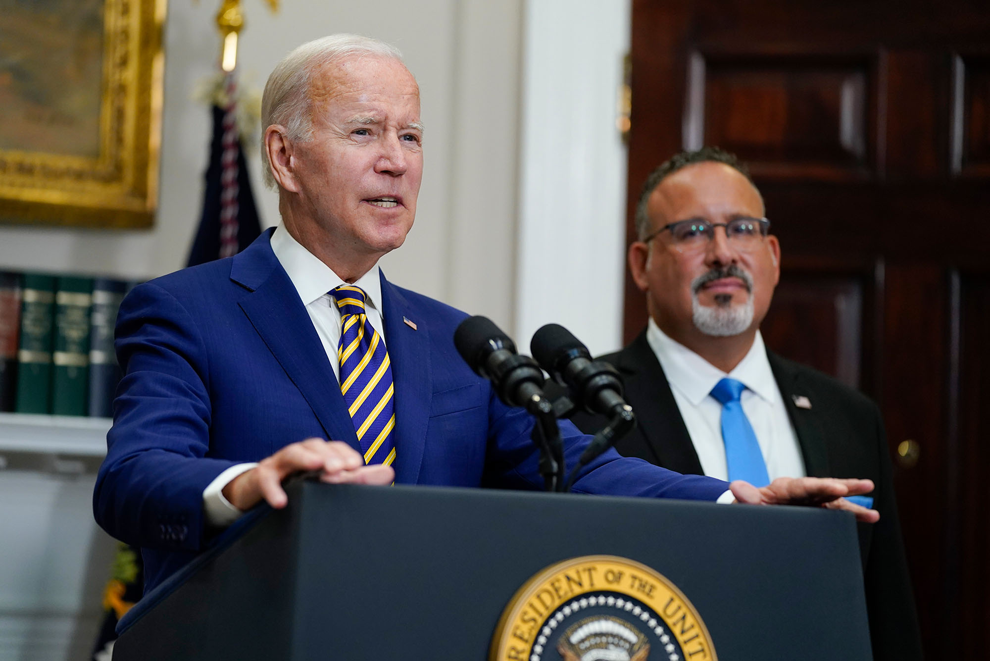 Photo of President Joe Biden speaking about student loan debt forgiveness in the Roosevelt Room of the White House on Wednesday, Aug. 24, 2022, in Washington. Education Secretary Miguel Cardona listens at right. An older white man wearing a navy suit and white collared shirt speaks into a microphone on a lectern. A Latino man wearing glasses, black blazer, and white collared shirt stands to his right.