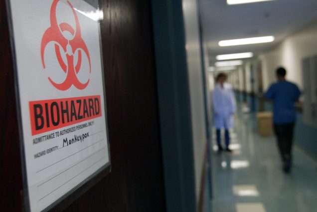 Photo of the University of Minnesota's Veterinary Diagnostic Laboratory door. A large sign with a red biohazard logo and the word "Biohazard" on it. It is seen in a fuzzy hall as two people wearing PPE walk.