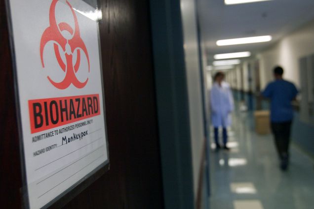 Photo of the University of Minnesota's Veterinary Diagnostic Laboratory door. A large sign with a red biohazard logo and the word "Biohazard" on it. It is seen in a fuzzy hall as two people wearing PPE walk.