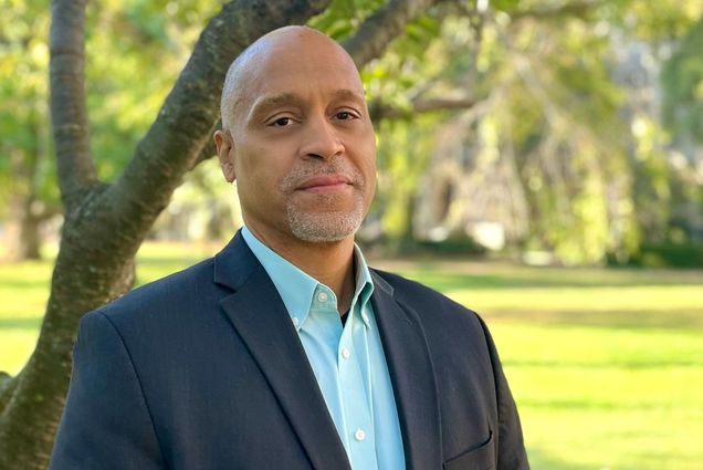 Photo of Scott Taylor. A bald black man with a neatly trimmed silver beard and mustache poses and looks directly at the camera. He wears a navy blazer and teal collared shirt and stands in front of a tree in a green field.