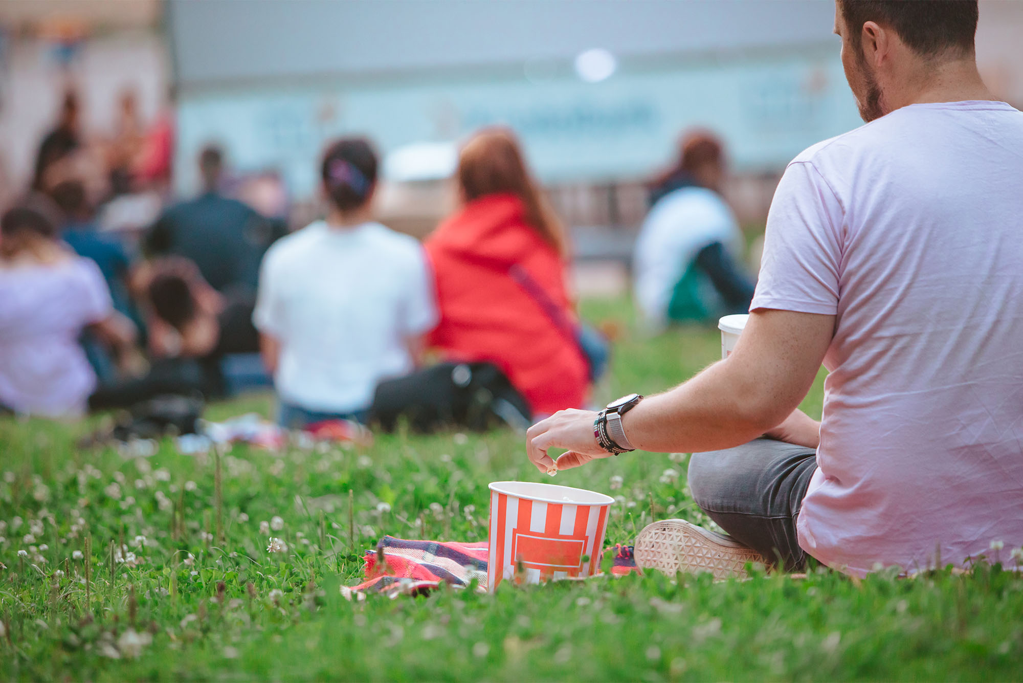A person sits on a grass lawn with a bucket of popcorn next to them, as other people sit nearby. All are looking at a large movie screen outdoors