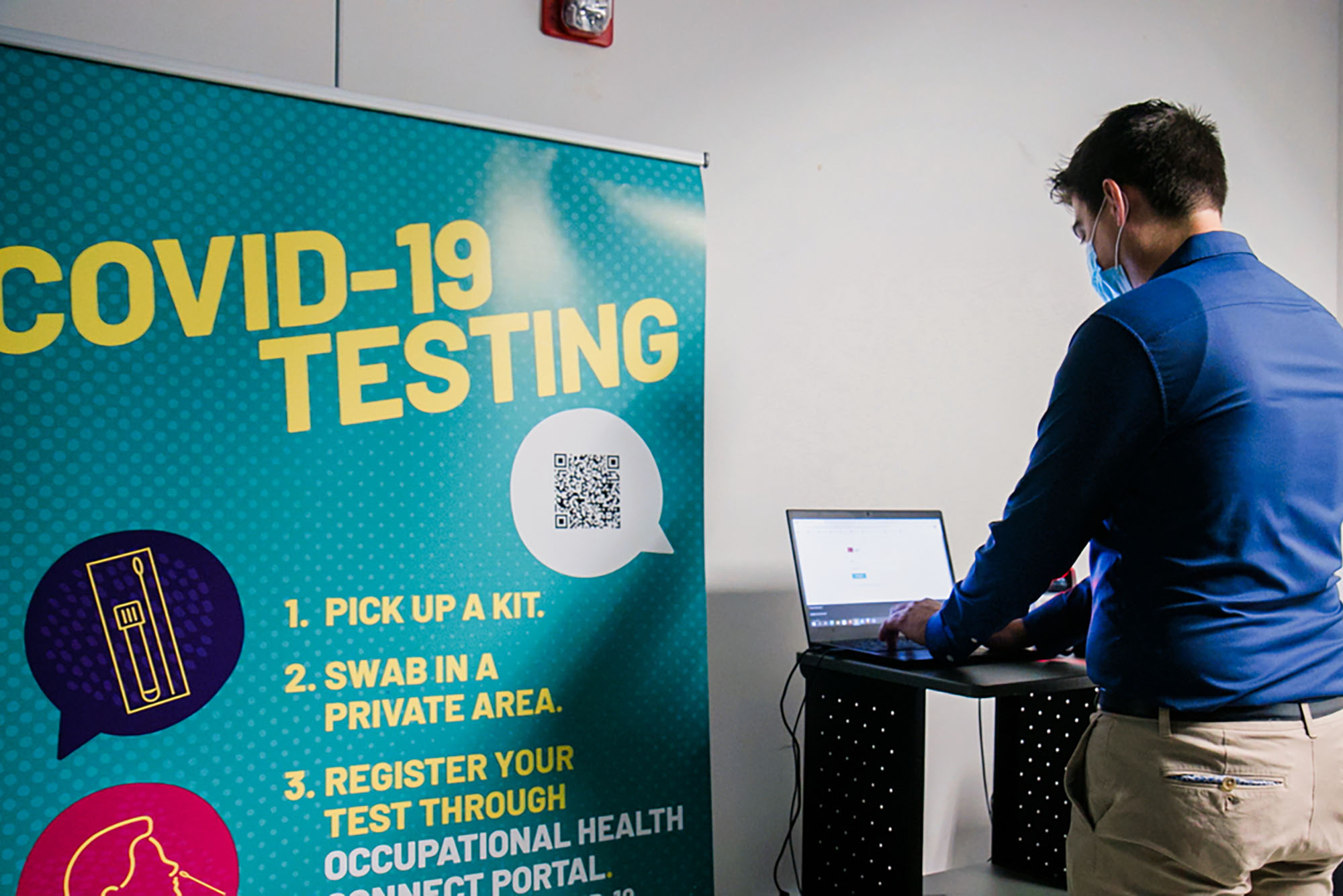 An individual working on a laptop stands with their back to the camera. A sign next to them reads "COVID-19 Testing" in yellow letters on a blue background