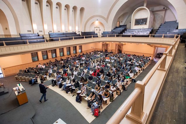 Wide photo of Uwe Beffert’s cell biology class September 2, 2021 in Morse Auditorium, when masking, but not social distancing, was required. The auditorium is two levels: the bottom floor has gray-blue chairs and carpeting, and the second level has black, wooden benches. The space is airy and light, and the bottom is filled with students holding notes and laptops on their laps as they look towards the front, where the lecturer, in a black jacket, is standing in front of a podium.