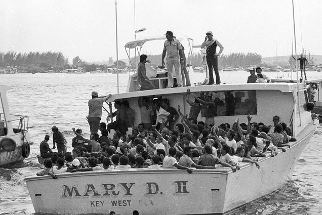 Black and white photo of a group of Cubans aboard the Mary D. II wave as they embark for U.S. shores from the port in Mariel Harbor, 20 miles west of Havana, Cuba, May 1980. The wide, white boat is jam-packed with folks, most of who look off to the left and raise their arms. Some stand on top of the boat near the helm. A shore with other boats is seen of in the distance.